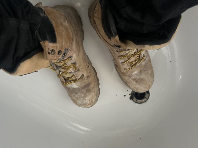 A top-down POV shot shows a person wearing scuffed and dirty tan work boots while standing in a white bathtub.