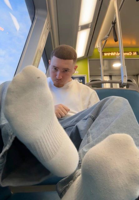 A young man wearing white socks and denim jeans sits on a public train.