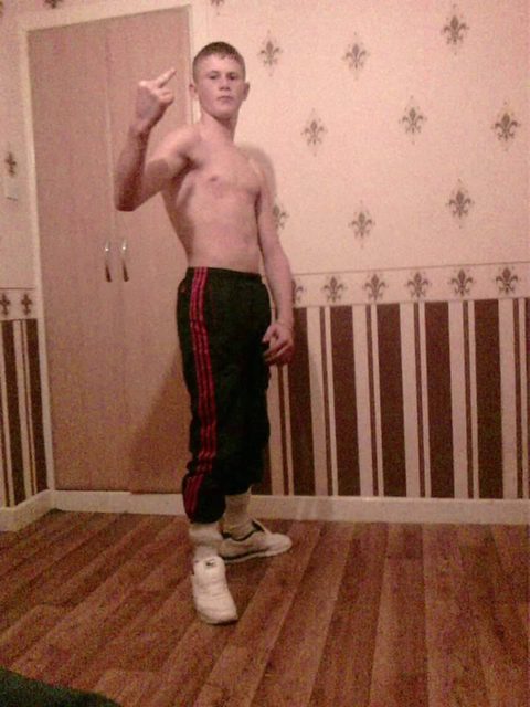 A shirtless young man with a buzzcut wears track pants tucked into socks and sneakers while posing indoors.