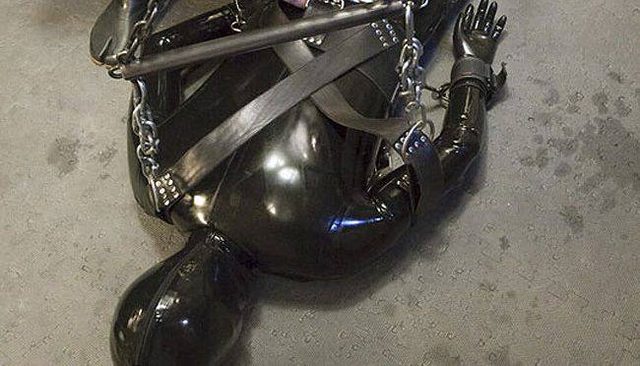 A person clad in a full black latex gimp suit and hood is bound on the floor with heavy straps, chains, cuffs, and a metal spreader bar.