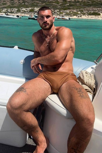 A muscular, hairy-chested man with tattoos and a beard sits outdoors on a boat in light brown swimsuit briefs.