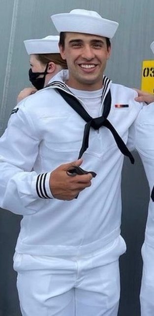 A young man in a white sailor uniform smiles, with another person partially visible in the background.