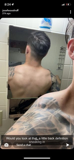 A man with tattoos is seen from behind, reflected in a bathroom mirror.