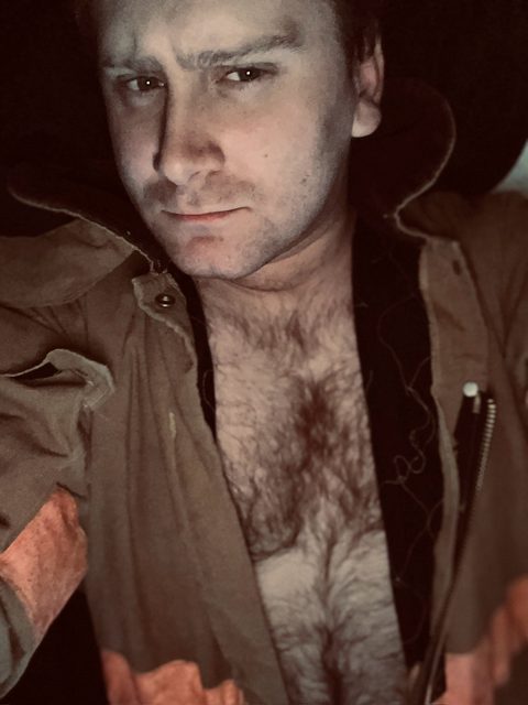 A man with a hairy chest wears an open jacket and looks at the camera.