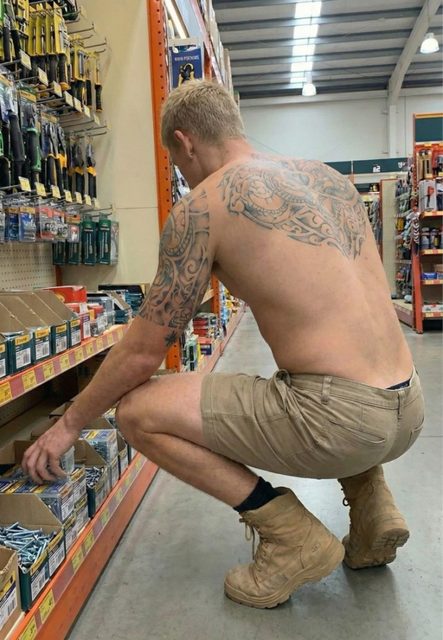 A shirtless, tattooed twunk with blonde hair crouches in a hardware store aisle wearing tan shorts and work boots.