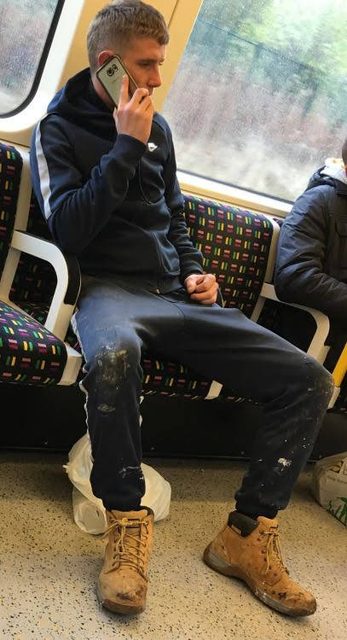 A man wearing a tracksuit and work boots sits on a train while talking on his phone.