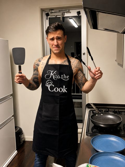 A shirtless, tattooed twunk stands in a kitchen wearing a black Kiss the Cook apron while holding a spatula and tongs.
