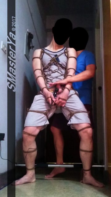 A man in a tank top and shorts is tied with rope while another man touches him in a bedroom.