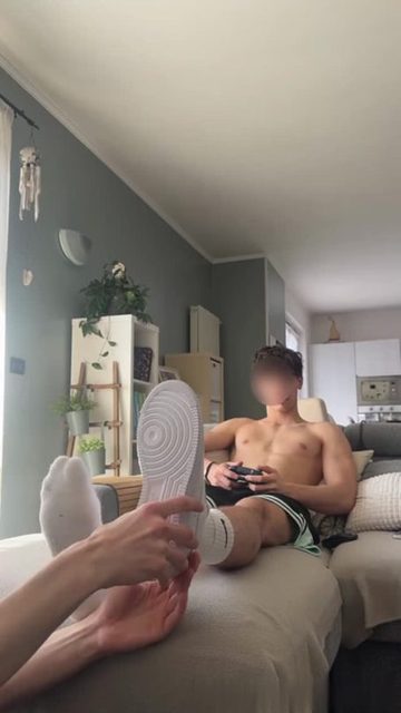 A shirtless man plays video games on a couch while another person touches his foot and sneaker.