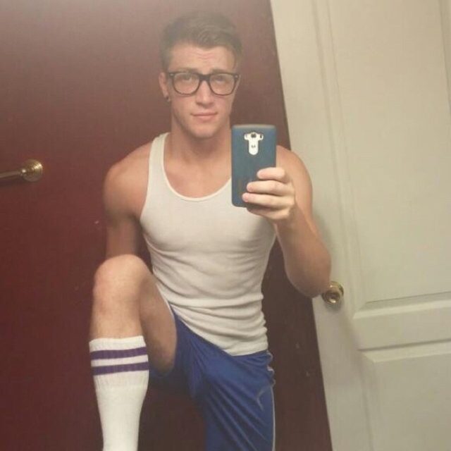 A young muscular man wearing a tank top, shorts, and tube socks takes a mirror selfie.