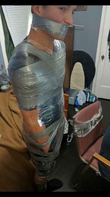 A young man is fully restrained with tape, including a mouth gag, in an indoor setting.