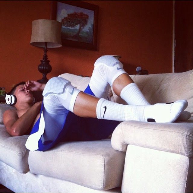 A shirtless athletic young man relaxes on a sofa wearing blue basketball shorts, padded knee sleeves, and white crew socks while listening to headphones.