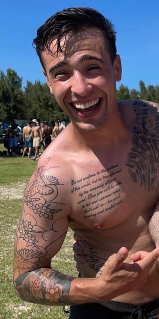 A smiling tattooed man poses shirtless at an outdoor public gathering.