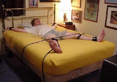 A man lies on a bed, restrained at his wrists and ankles by ropes and tape.