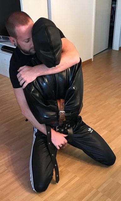 One man embraces another man who is in a latex straitjacket and mask, kneeling on a wooden floor.