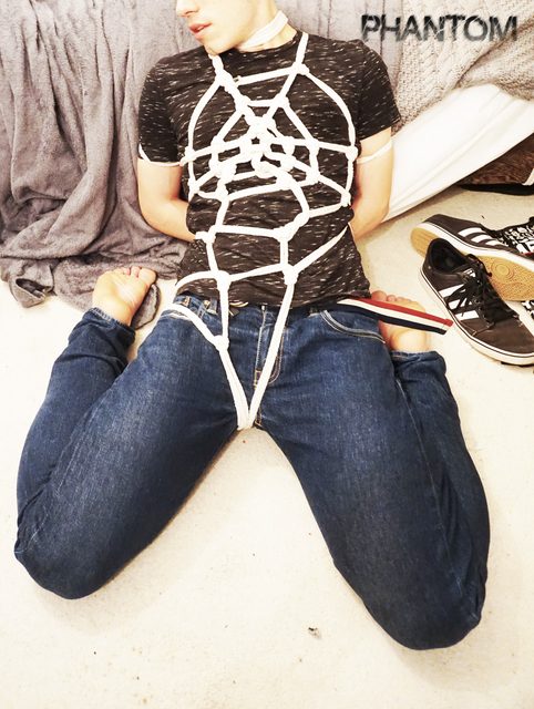 A young man tied in a detailed white rope harness over his t-shirt and jeans sits kneeling on the floor with his hands bound behind his back.