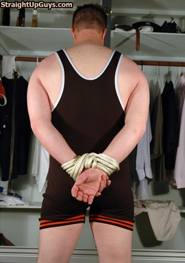 Rear view of a man in a wrestling singlet standing in front of an open closet with his hands bound behind his back with thick rope.