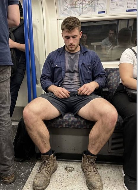 A muscular man with thick, hairy legs and a sweaty t-shirt sits with his legs spread wide on a subway train.