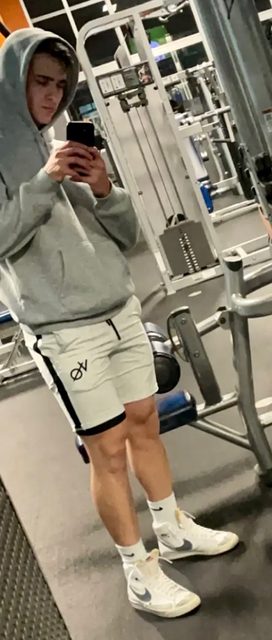 A young man in a hoodie and shorts takes a mirror selfie in a gym.