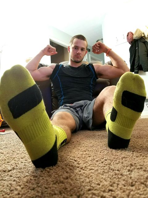 A muscular man in sportswear and athletic socks flexes his biceps while sitting on a carpeted floor.