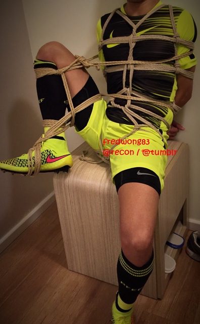 A man in sportswear is tied with rope bondage while sitting indoors.