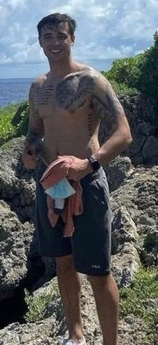 A smiling muscular man with tattoos stands shirtless on a rocky outdoor landscape by the ocean.