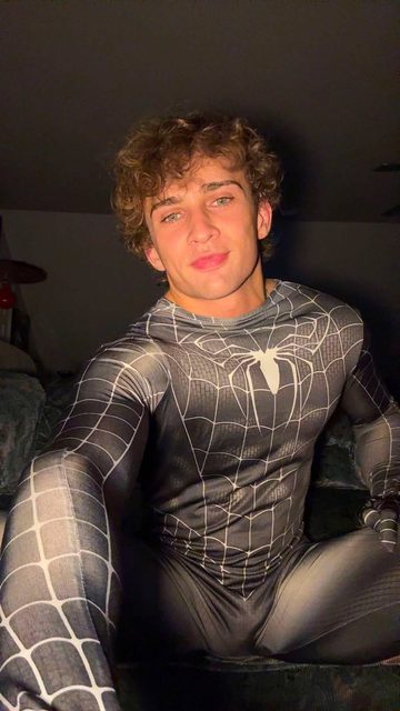 A young muscular man with curly hair poses in a black Spider-Man costume.