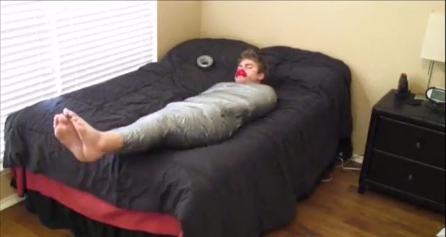 A man is fully restrained with tape on a bed in a bedroom.