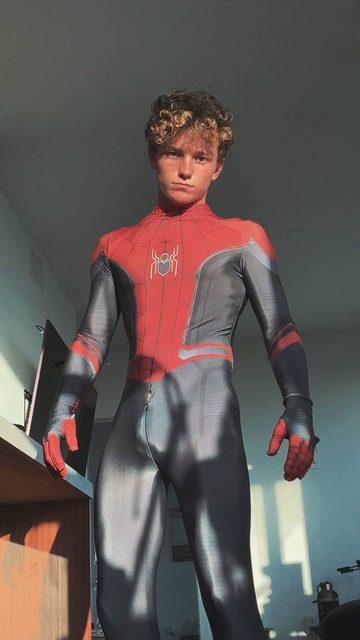 A young man stands indoors wearing a form-fitting Spider-Man costume with a front zipper.