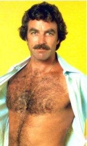 A mature man with a thick mustache displays his hairy chest through an open light blue shirt in front of a yellow backdrop.