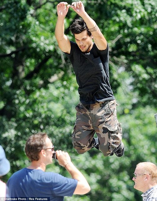 A muscular young man wearing camouflage cargo pants and a black t-shirt is captured mid-air during a jump outdoors.