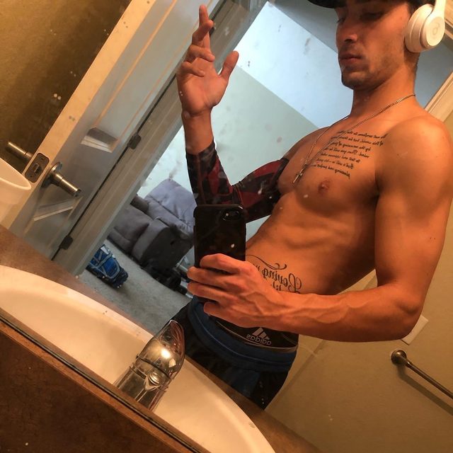 A muscular man takes a mirror selfie in a bathroom, wearing a cap and headphones.