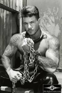 A black and white photograph of a muscular, tattooed man wearing a leather collar and leather pants while holding heavy metal chains.