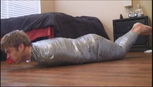 A man is fully restrained with duct tape and a gag while lying on a wooden floor in a bedroom.
