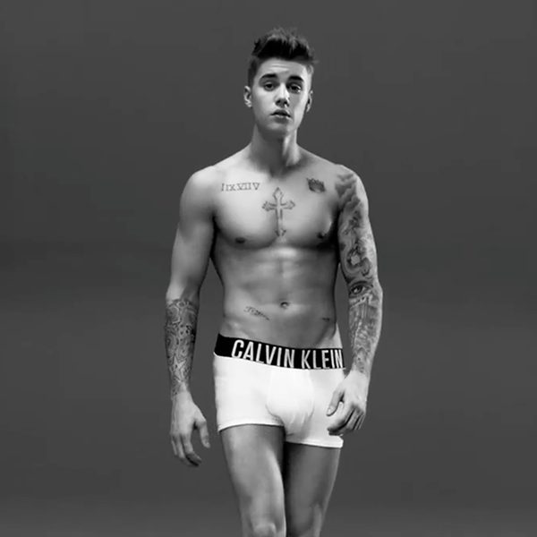 A lean, tattooed young man poses in white Calvin Klein boxer briefs against a plain studio background.