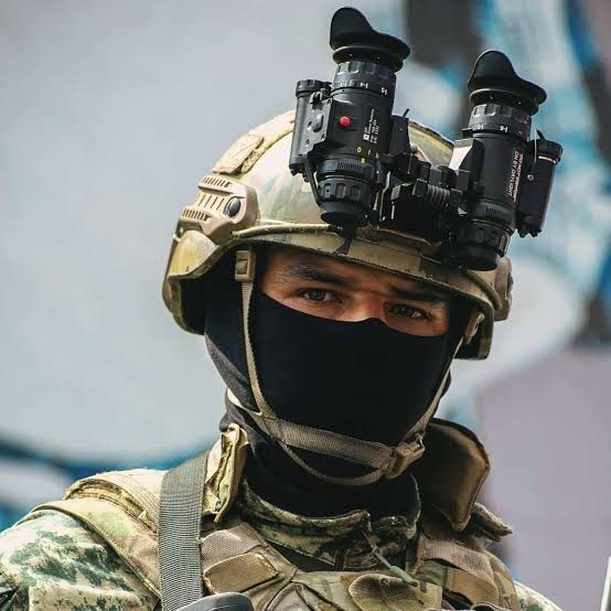 A man in tactical military gear wears a camouflage uniform, a helmet with night vision goggles, and a black balaclava.