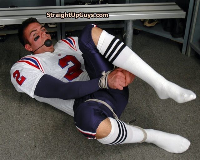 A man dressed in a football uniform lies bound with rope and gagged with a ball gag on a locker room floor.