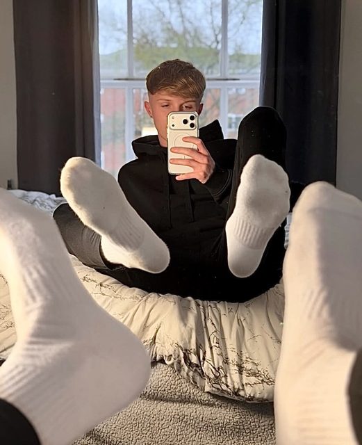 A young man takes a selfie while lounging on a bed, showing off his white socks.