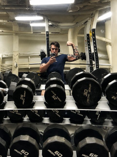 A muscular man with arm tattoos flexes his bicep for a mirror selfie in a weight room.