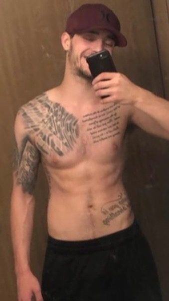 A muscular man with tattoos and a cap takes a mirror selfie.