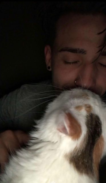 A man with piercings cuddles a cat in a dimly lit room.