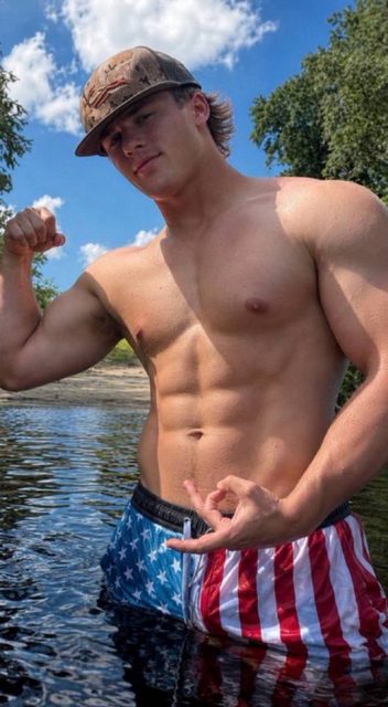 A shirtless, muscular young man flexes his bicep while standing in a lake wearing American flag board shorts and a camo cap.