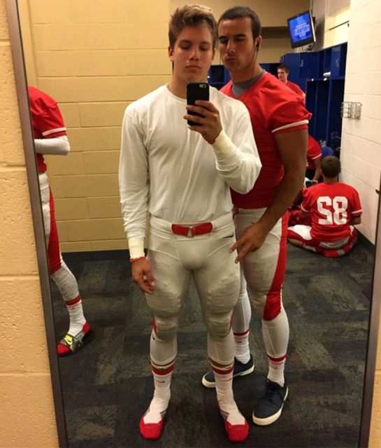 Two athletic young men pose for a mirror selfie inside a locker room wearing tight football kits and compression gear.