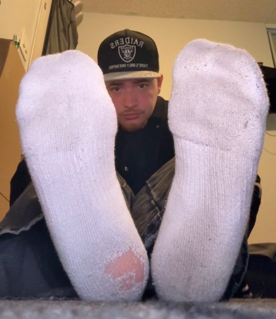 A young man wearing a cap and beard poses with his dirty and ripped socks facing the camera.