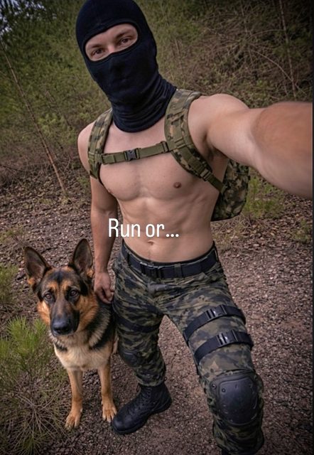 A shirtless muscular man wearing a balaclava and camo tactical gear stands with a German Shepherd in a wooded area.