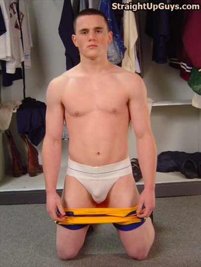 A muscular young man with a buzzcut kneels in a locker room wearing only a white jockstrap while holding his athletic shorts down.