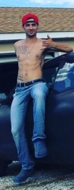 A shirtless man in jeans and a red cap leans on a car outdoors.