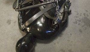 A person fully encased in a shiny black latex suit lies bound on a concrete floor with heavy metal chains and bondage straps.