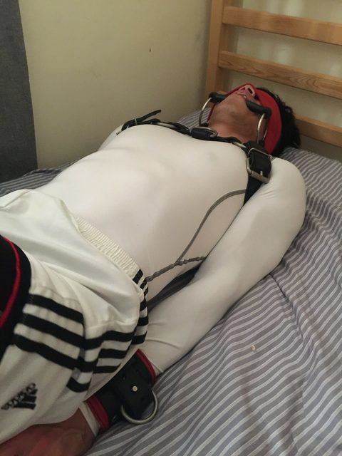 A man lies restrained in a bed, wearing a blindfold, bit gag, harness, and sportswear.