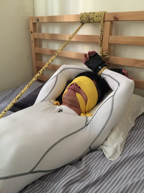 A man is tied to a bed headboard with rope and blindfolded in a bedroom.